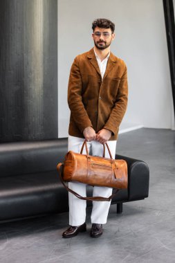 Kahverengi süet ceketli ve beyaz pantolonlu yakışıklı bir adam modern iç mekanda özel deri çanta taşıyor..