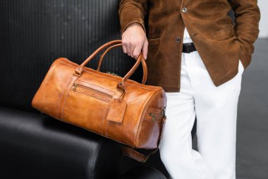Süet ceket ve beyaz pantolon giyen, deri bir spor çantası tutan bir adamı yakından çek..