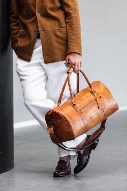 Süet ceket ve beyaz pantolon giyen kahverengi spor çantalı bir adamın yakın plan fotoğrafı..