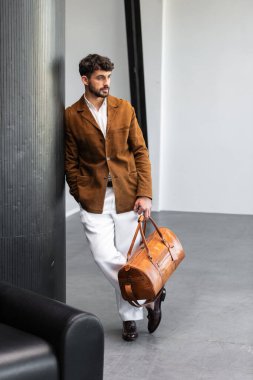 Kahverengi süet ceketli ve beyaz pantolonlu yakışıklı bir adam modern iç mekanda özel deri çanta taşıyor..