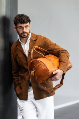 Kahverengi süet ceketli ve gözlüklü şık bir adam elinde deri bir çantayla bir sütuna yaslanıyor..