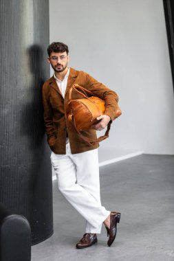 Kahverengi süet ceketli ve beyaz pantolonlu yakışıklı bir adam modern iç mekanda özel deri çanta taşıyor..