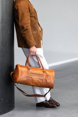 Süet ceket ve beyaz pantolon giyen kahverengi spor çantalı bir adamın yakın plan fotoğrafı..