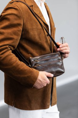 Kahverengi süet ceketli, beyaz pantolonlu şık bir adam. Duvara yaslanmış, elinde küçük, siyah deri bir çanta tutuyor..