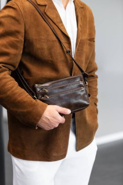 Kahverengi süet ceketli, beyaz pantolonlu şık bir adam. Duvara yaslanmış, elinde küçük, siyah deri bir çanta tutuyor..