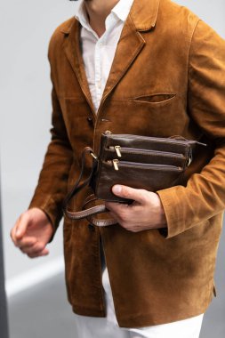Kahverengi süet ceketli, beyaz pantolonlu şık bir adam. Duvara yaslanmış, elinde küçük, siyah deri bir çanta tutuyor..