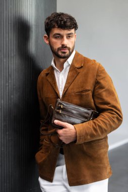 Kahverengi süet ceketli, beyaz pantolonlu şık bir adam. Duvara yaslanmış, elinde küçük, siyah deri bir çanta tutuyor..