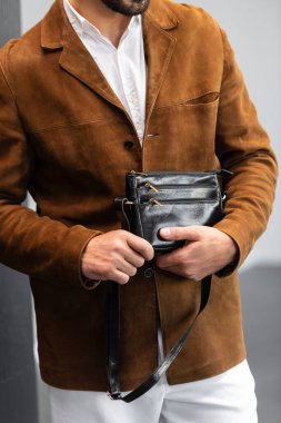 Kahverengi süet ceketli, beyaz pantolonlu şık bir adam. Duvara yaslanmış, elinde küçük, siyah deri bir çanta tutuyor..