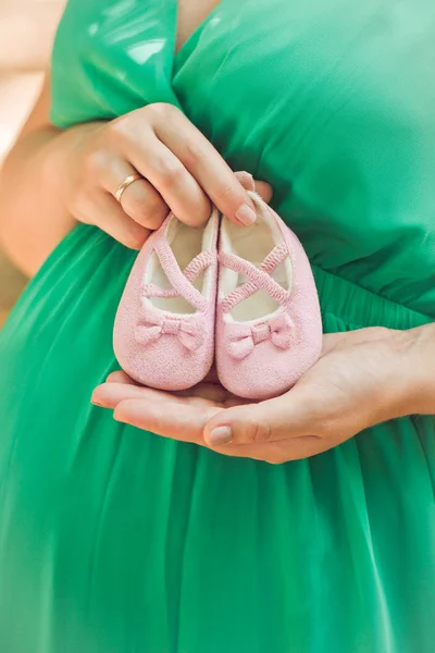 Embarazada mujer en verde vestido vientre celebración rosa bebé botines, e — Foto de Stock