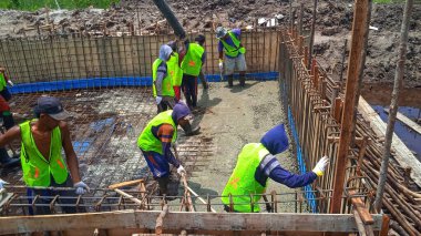 Endonezyalı veya Güneydoğu Asyalı inşaat işçileri beton dökerek, biçimlendirmenin içindeki taze çimento karışımını çelik takviyesi (inşaat demiri) ile dengeliyor.