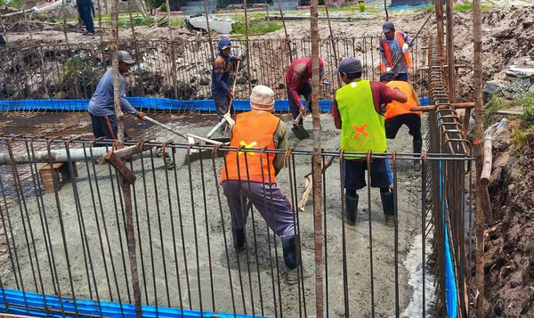 Endonezyalı veya Güneydoğu Asyalı inşaat işçileri beton dökerek, biçimlendirmenin içindeki taze çimento karışımını çelik takviyesi (inşaat demiri) ile dengeliyor.