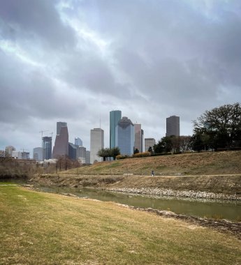 Buffalo Bayou Parkı 'nın bulutlu manzarasında Teksas şehir merkezindeki binalar ve ağaçlar çimlerle kaplı.