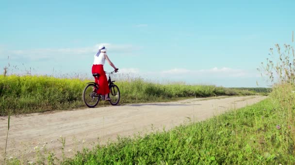 Signora in bicicletta in bianco cappello cavalca sul campo su una strada di campagna lontano dalla fotocamera — Video Stock