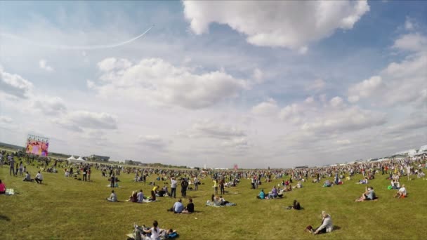 ЖУКОВСКИЙ, РОССИЯ - 27 АУГ: Много людей отдыхают в летний день в поле и смотрят авиашоу. Самолеты летают над аэродромом на авиашоу с белым дымом — стоковое видео