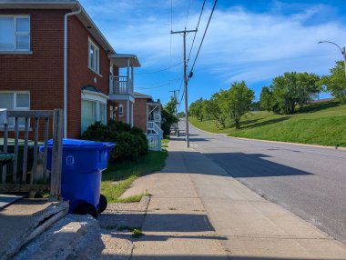 Kanada, 09 Temmuz 2025: Boş güneşli caddenin yanında mavi geri dönüşüm kutulu kırmızı tuğlalı evler