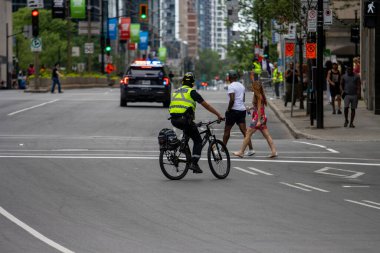 Kanada, 05 Temmuz 2025: Bisikletli polis memuru yayaları şehir caddesinde karşıdan karşıya geçerken izliyor