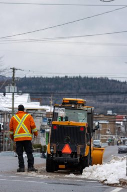 Kanada, Quebec, 27 Kasım 2025: Parlak ceketli işçi kar küreme aracının buzlu yolu temizlemesine rehberlik ediyor