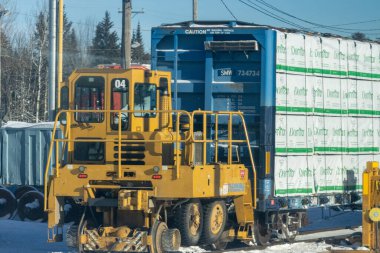 MONTREAL, CANADA, 26 Aralık 2025: Sarı şalter lokomotifi, mavi yük arabası, istiflenmiş endüstriyel malzemeler, karla kaplı zemin, elektrik direkleri ve kabloları