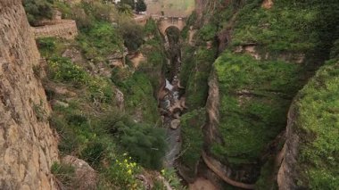 Alçak yeşil El Tajo Kanyonu 'na bakan sinematik yüksek açılı bir çekim. Görüntüler nehrin dikey uçurumlar arasında Puente Viejo taş köprüsünün odak noktası olarak aktığını gösteriyor..