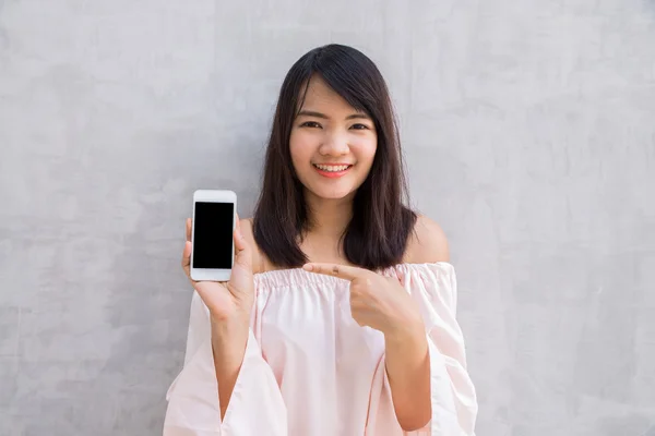 Hymyilevä nainen osoittaa tyhjä älypuhelin näyttö seisoo betoniseinällä . — kuvapankkivalokuva