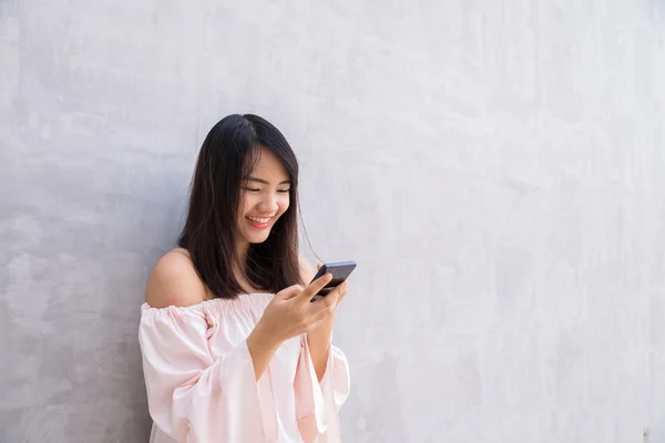 Kaunis aasialainen nainen käyttää matkapuhelinta, betoniseinän yli — kuvapankkivalokuva