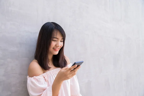 Kaunis aasialainen nainen käyttää matkapuhelinta, betoniseinän yli — kuvapankkivalokuva