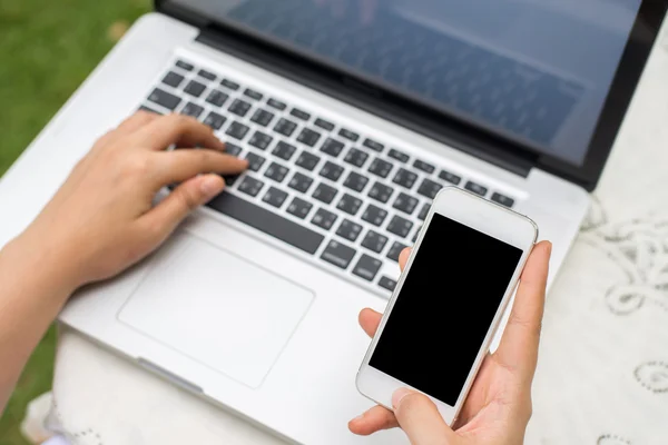 Telefone celular com tela em branco em mãos de mulher e laptop na mesa de madeira, zombar — Fotografia de Stock