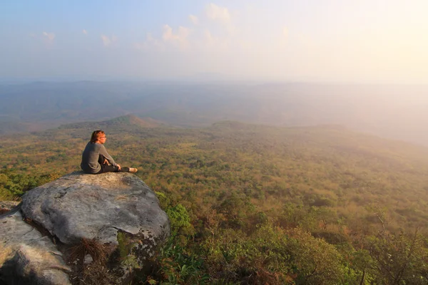 Adult tourist in black trousers, jacket and dark cap sit on cliff's edge and looking to misty hilly valley bellow — Stock Photo, Image