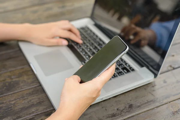 Mobiltelefon med blank skærm i kvindens hænder og laptop på træbord, mock up - Stock-foto