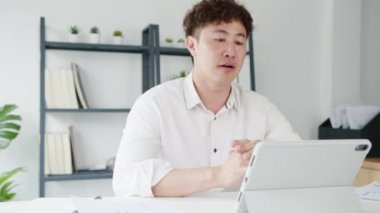 Tablet kullanan genç Asya 'lı işadamı, evdeki oturma odasında akıllı bir şekilde çalışırken iş arkadaşlarıyla görüntülü görüşme planı hakkında konuşuyor. Kişisel izolasyon, sosyal uzaklık, korona virüsünün önlenmesi için karantina.