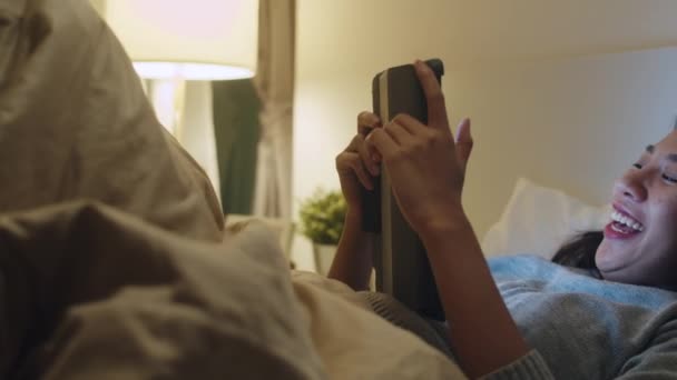 年轻的手机成瘾的亚洲女士使用平板电脑检查社交媒体 晚上睡觉前在床上开心的笑着 漂亮迷人的女孩笑着在卧室里放松一下 — 图库视频影像