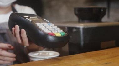 Genç Asyalı bayan barista, yüz maskesi takıyor. Sıcak kahve fincanını tüketiciye götürüyor ve kafede kredi kartı kullanıyor. Corona virüsü ve sosyal uzaklıktan sonra yaşam tarzı yeni normal.
