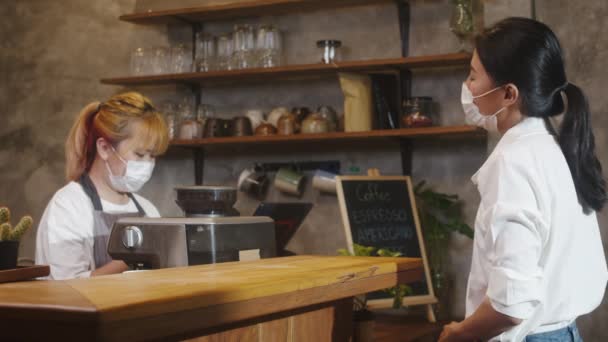 若いアジアの女性バリスタは フェイスマスクを着用し ホットコーヒーペーパーカップを消費者に持ち去り カフェでの支払いにクレジットカードを使用します コロナウイルスと社会的距離の後のライフスタイルの新しい正常な — ストック動画