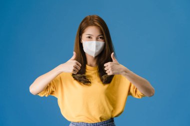 Tıbbi yüz maskesi takan genç Asyalı kız başparmağına günlük giysiler giydirmiş ve mavi arka planda izole edilmiş kameraya bakıyor. Kişisel izolasyon, sosyal uzaklık, korona virüsü için karantina.