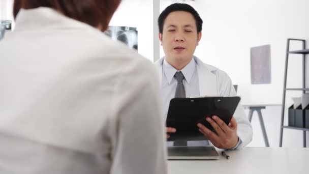 クリップボードを使用して白い医療制服の深刻なアジアの男性医師は素晴らしいニューストークの結果や症状を提供しています健康クリニックや病院のオフィスでデスクに座って女性患者と議論 — ストック動画