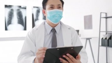 Ciddi Asyalı erkek doktor, pano kullanarak koruyucu maske takıyor. Harika haberler sunuyor. Sonuçları ya da kadın hastalarla hastane ofisinde belirtileri tartışıyor. Corona virüsünden sonra yaşam tarzı yeni normal.