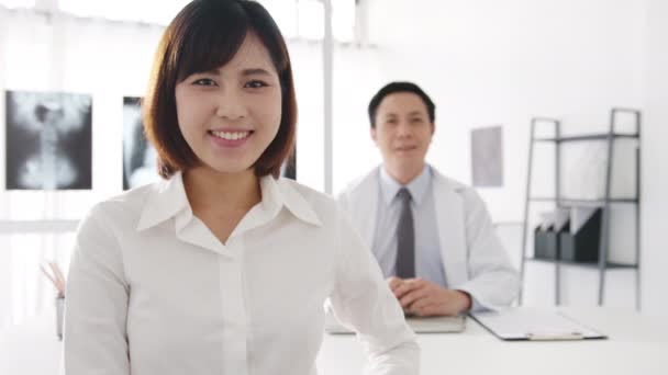 有信心的亚洲男性医生身穿白衣 年轻的女病人在诊所或医院的办公桌前进行医疗会诊时 看着相机和微笑 咨询和治疗概念 — 图库视频影像