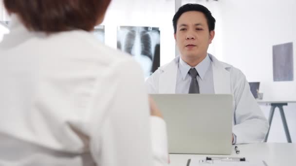コンピュータのラップトップを使用して白い医療制服を着た深刻なアジアの男性医師は素晴らしいニューストークの結果や症状を提供しています女性患者との議論健康クリニックや病院のオフィスの机に座って — ストック動画