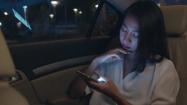 Moda bürosu kıyafetleri içinde başarılı genç Asyalı iş kadını gece geç saatlere kadar şehir modern şehrinde arabanın arka koltuğunda otururken akıllı telefon kullanıyor. İnsanların mesleki tükenme sendromu kavramı.