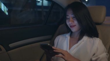 Moda bürosu kıyafetleri içinde başarılı genç Asyalı iş kadını gece geç saatlere kadar şehir modern şehrinde arabanın arka koltuğunda otururken akıllı telefon kullanıyor. İnsanların mesleki tükenme sendromu kavramı.