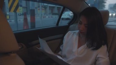 Moda ofis kıyafetleri içinde başarılı genç Asyalı iş kadını sabahın erken saatlerinde şehir merkezinde arabanın arka koltuğunda çalışıyor. İş konseptiyle ilgili işler.