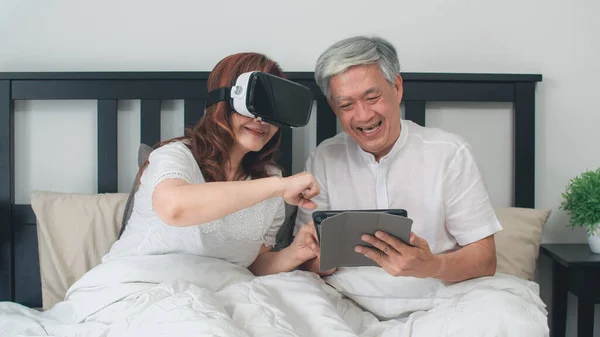 Asian senior couple using tablet at home. Asian Senior Chinese grandparents, feeling happy fun and VR playing games together while lying on bed in bedroom at home in the morning concept.