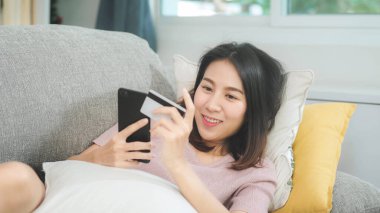 Genç gülümseyen Asyalı kadın evde oturma odasında dinlenirken kredi kartıyla online alışveriş yapıyor. Ev konseptinde Latin ve Latin kökenli kadınlar var..