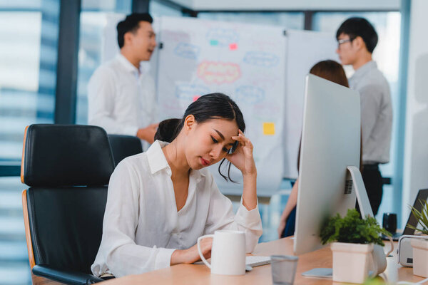 Тысячелетняя молодая китайская предпринимательница работает над проблемой исследования проекта на рабочем столе компьютера в конференц-зале в маленьком современном офисе. Концепция синдрома профессионального выгорания.