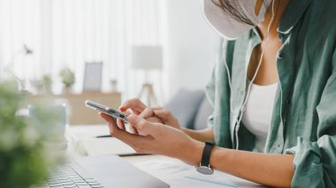 Serbest çalışan Asyalı kadınlar oturma odasında masa başında otururken internet üzerinden akıllı telefon alışverişi yaparak yüz maskesi takıyorlar. Evden çalışmak, uzaktan çalışmak, sosyal mesafeler, koronavirüs için karantina.