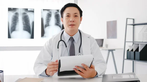 Young Asia male doctor in white medical uniform with stethoscope using computer laptop talk video conference call with patient, looking at camera in health hospital. Consulting and therapy concept.