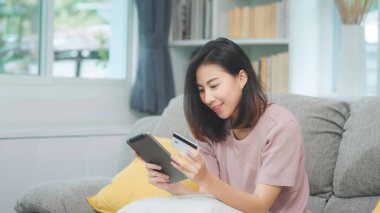 Genç gülümseyen Asyalı kadın evde oturma odasında dinlenirken kredi kartıyla online alışveriş yapıyor. Ev konseptinde Latin ve Latin kökenli kadınlar var..