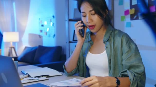 Szabadúszó ázsiai nők segítségével laptop beszélni telefonon elfoglalt vállalkozó távolról dolgozik a nappaliban. Éjszakai háztúlterhelés, távoli munka, társadalmi távolság, karantén a koronavírus miatt..