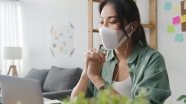 Bilgisayarı kullanarak tıbbi maske takan Asyalı iş kadını, evdeki oturma odasında çalışırken iş arkadaşlarıyla görüntülü görüşme planı hakkında konuşuyor. Corona virüsünün önlenmesi için sosyal mesafe ve karantina.