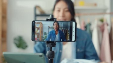 Genç Asyalı bayan moda tasarımcısı cep telefonu siparişi alıyor ve mağazada canlı yayın yapan kıyafetleri gösteriyor. Küçük işletme sahibi, çevrimiçi pazar dağıtımı kavramı.